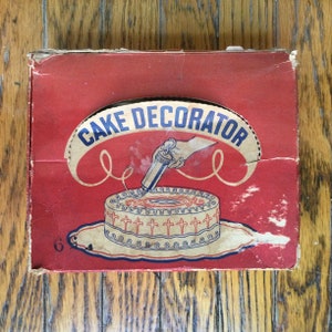 May include: Vintage red and white cardboard box with the text "Cake Decorator" in a white circle. The box features an illustration of a cake with white frosting and a piping bag decorating the cake.