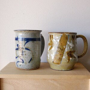 Puede incluir: Dos tazas de cer&aacute;mica hechas a mano. La taza de la izquierda es azul claro con dise&ntilde;os abstractos azul oscuro y un asa. La taza de la derecha es de color canela con dise&ntilde;os abstractos marrones y blancos y un asa. Ambas tazas est&aacute;n sobre una superficie de madera.