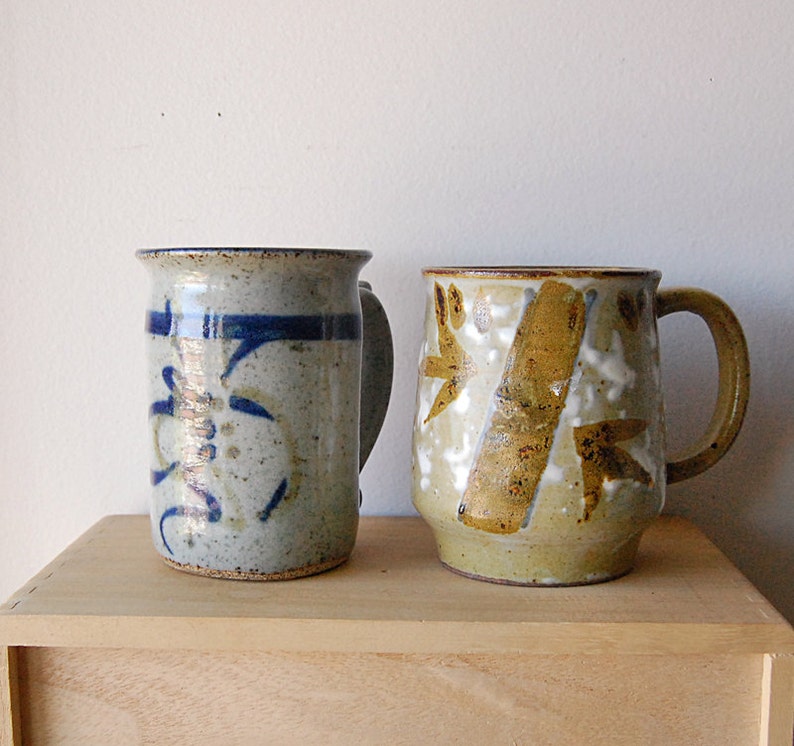 Puede incluir: Dos tazas de cer&aacute;mica hechas a mano. La taza de la izquierda es azul claro con dise&ntilde;os abstractos azul oscuro y un asa. La taza de la derecha es de color canela con dise&ntilde;os abstractos marrones y blancos y un asa. Ambas tazas est&aacute;n sobre una superficie de madera.