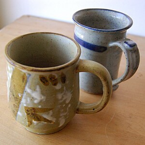 Puede incluir: Dos tazas de cer&aacute;mica hechas a mano. La taza delantera es de color verde oliva con detalles blancos y marrones. La segunda taza es azul claro con una franja y asa azul oscuro. Ambas tazas tienen un aspecto r&uacute;stico y texturizado, sobre una superficie de madera.