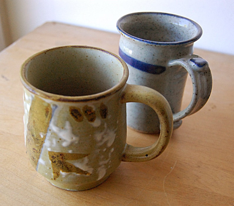 Puede incluir: Dos tazas de cer&aacute;mica hechas a mano. La taza delantera es de color verde oliva con detalles blancos y marrones. La segunda taza es azul claro con una franja y asa azul oscuro. Ambas tazas tienen un aspecto r&uacute;stico y texturizado, sobre una superficie de madera.