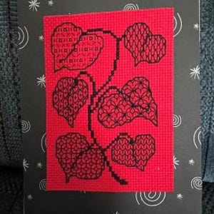May include: A red cross-stitch card with a black design of a stylized plant with leaves. The leaves have a geometric pattern.