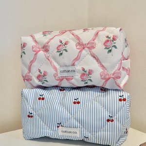 May include: Two quilted makeup bags, one with a pink and white floral pattern and the other with a blue and white striped pattern. Both bags have a white tag with the text "cotton co."