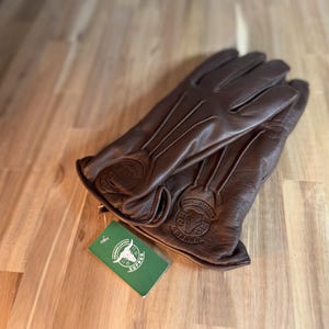 May include: Brown leather gloves with a stitched design and a bull logo on the cuff. The gloves are folded in half and a green tag with a bull logo and the word "SUPREME" is visible.
