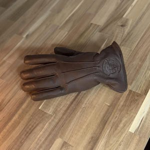 May include: Brown leather gloves with a stitched design and a logo on the back of the hand. The logo is a circle with a skull inside and the text "Iron & Resin" around it.