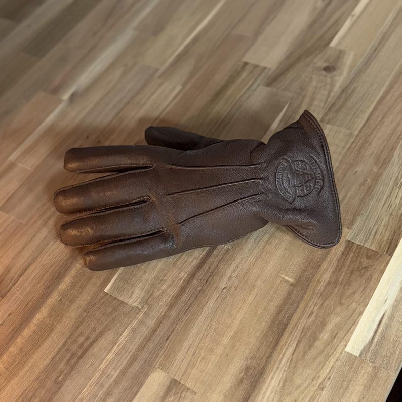 May include: Brown leather gloves with a stitched design and a logo on the back of the hand. The logo is a circle with a skull inside and the text "Iron & Resin" around it.