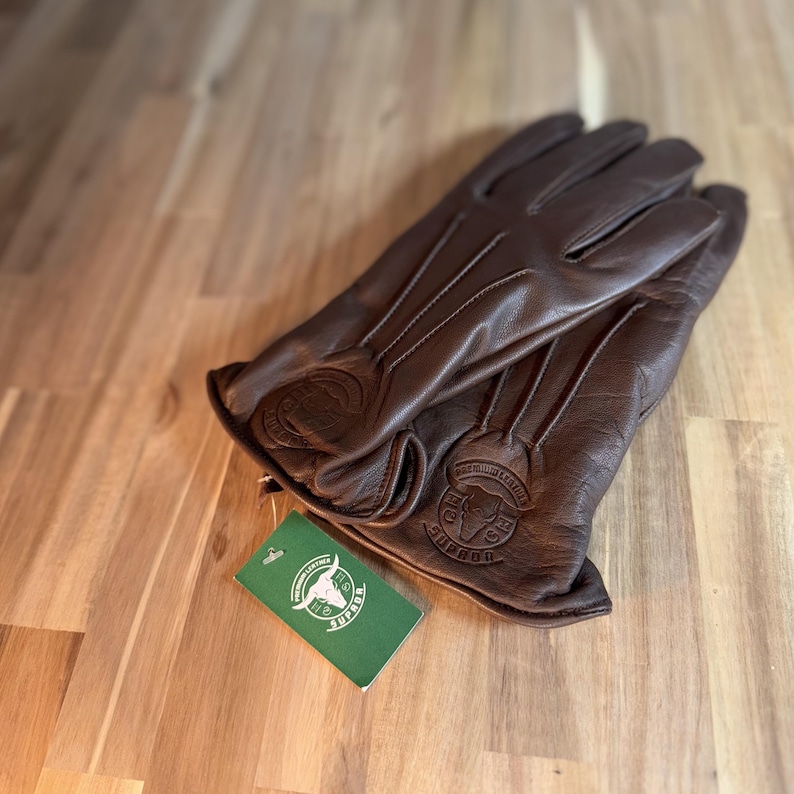 May include: Brown leather gloves with a stitched design and a bull logo on the cuff. The gloves are folded in half and a green tag with a bull logo and the word "SUPREME" is visible.