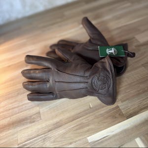 May include: A pair of brown leather gloves with a logo on the back of the hand. The logo is a stylized image of a dog's head with the text "The Dog's Bollocks" underneath.