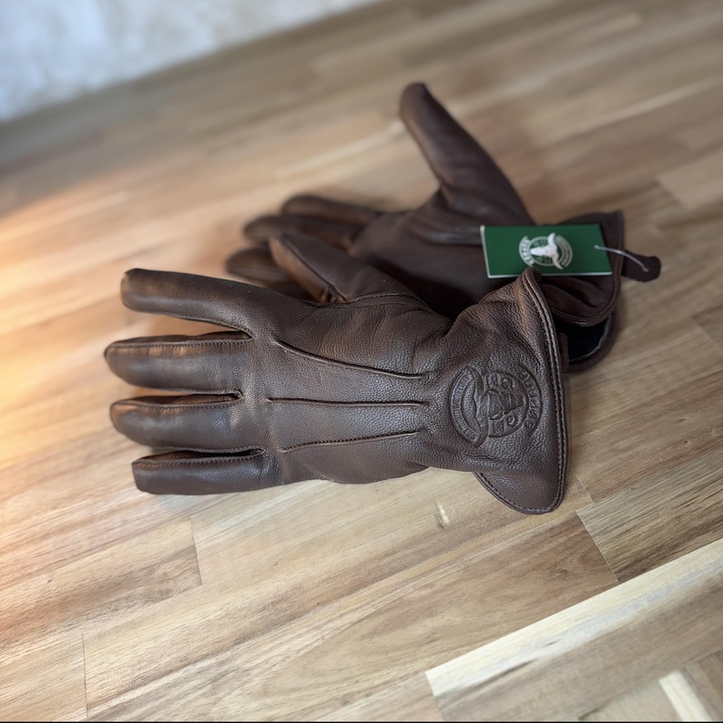 May include: A pair of brown leather gloves with a logo on the back of the hand. The logo is a stylized image of a dog's head with the text "The Dog's Bollocks" underneath.