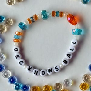 May include: A beaded bracelet with the words "OKC KNOWS BALL" spelled out in white letter beads. The bracelet features blue, orange, and clear beads. Additional beads in various colors surround the bracelet.