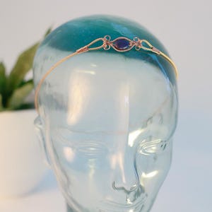 May include: A delicate copper wire headband with a central oval-shaped, dark blue gemstone. The wire is intricately woven around the gemstone and at the sides, creating a decorative accent. The headband is displayed on a glass head.