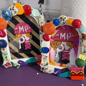 May include: Two decorative picture frames with colorful embellishments. Each frame features a cartoon image with the text "MP PINK" and is surrounded by various decorative elements, including beads and painted shapes.