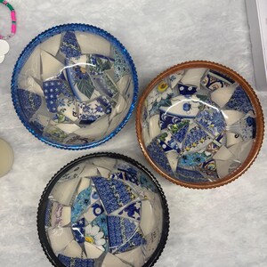 May include: Three mosaic bowls with blue, brown, and black rims. Each bowl is filled with broken pieces of blue and white patterned china. The china pieces have floral and geometric designs. The bowls are on a light grey surface.