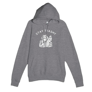 May include: A grey hooded sweatshirt with a kangaroo pocket and drawstring. The front features a white graphic of a raccoon in a bin with the words "STAY TRASHY" above it. The hoodie has a hood and long sleeves.
