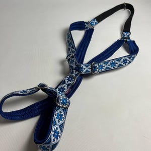 May include: A blue dog harness featuring a white and light blue geometric pattern on the straps. The harness includes silver-coloured metal hardware, adjustable sliders, and a black elastic section at the top, designed with a multi-strap configuration.
