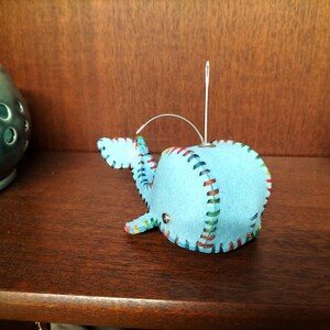 Może przedstawiać: Jasnoniebieska filcowa ozdoba w kształcie wieloryba z kolorowymi, szytymi krawędziami. Wieloryb ma ogon i płetwy, a igła jest przymocowana do zawieszenia. Ozdoba znajduje się na ciemnej drewnianej powierzchni.