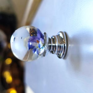 May include: A decorative cabinet knob with a clear, spherical glass head. The head encases a blue and white floral arrangement. The knob has a shiny, silver-colored metal base with a ridged design. The knob is mounted on a white surface.