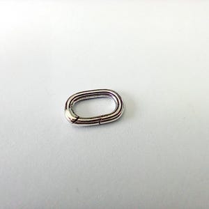 Può includere: Un anello ovale color argento, probabilmente per la creazione di gioielli o artigianato, è mostrato su uno sfondo bianco. L'anello ha un design a cerniera per una facile apertura e chiusura. Il metallo ha una superficie lucida e riflettente.