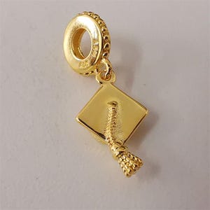May include: A gold-colored charm featuring a graduation cap design. The charm has a square cap with a tassel and a ring for attachment. The charm is made of a shiny, metallic material.