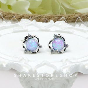 May include: A pair of silver-toned stud earrings. Each earring features a light purple opal-like sphere, surrounded by a swirling silver design and small clear accent stones. The earrings are displayed on a white decorative surface. The text "SHARESTORESHOP Handcrafted Jeweller" is visible.