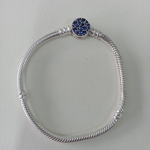 May include: A silver bracelet with a snake chain design. The bracelet features a round charm set with numerous small, dark blue stones. The charm is centered on the bracelet, adding a touch of elegance to the piece.
