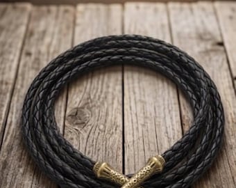 May include: A black braided cord necklace or belt featuring two crossed, textured brass-toned metal tips at the ends. The cord is coiled into a circular shape, showcasing a consistent woven pattern throughout the length of the dark material.