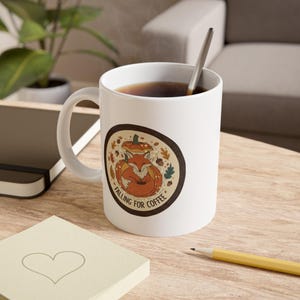 May include: White ceramic coffee mug with a "Falling for Coffee" design. The design features a fox curled up inside a pumpkin, surrounded by autumn leaves and acorns. A silver spoon rests inside the mug, which is filled with coffee.