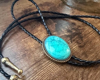 May include: A bolo tie featuring a large, oval-shaped turquoise stone with dark veining, set in a textured brass-toned metal frame. The cord is made of black braided material, finished with two matching brass-toned metal tips with engraved details.
