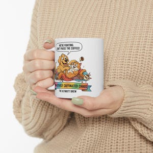 Op de afbeelding: Witte keramische mok met een cartoon illustratie van een hond en een kat. De mok heeft de tekst "WE'RE FIGHTING, DON'T PAUSE THE COFFEE!" en "POSITIVELY CAFFINATED CHAOS THE ULTIMUTT BREW".