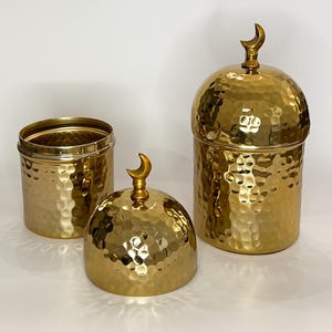 以下が含まれることがあります： 金色の金属製の容器が2つあり、ハンマーで叩いたような仕上げと三日月形の飾りがついています。大きな容器には蓋があり、小さな容器は開いています。