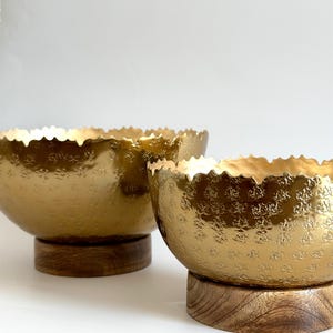 May include: Two gold-colored decorative bowls with textured surfaces and scalloped rims. The bowls sit on round, wooden bases. The background is a plain white.
