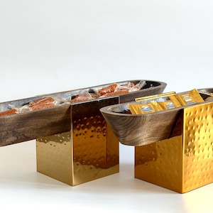 Può includere: Due ciotole di legno scuro su supporti in metallo dorato strutturato. Una è lunga e rettangolare, piena di articoli confezionati singolarmente. L'altra è più rotonda e contiene piccoli pacchetti.