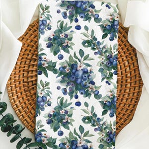 May include: A white fabric with a repeating pattern of blue blueberries, green leaves, and white flowers. The fabric is on a woven brown basket.