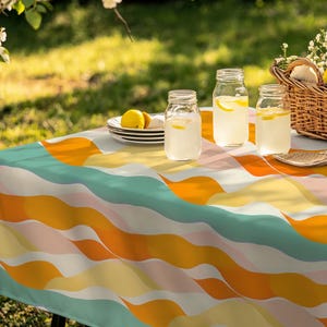 May include: A colorful outdoor picnic scene with a vibrant tablecloth featuring a wavy pattern in shades of orange, teal, yellow, and pink. Three glass jars with lemonade and lemon slices sit on the table, along with a basket and plates.