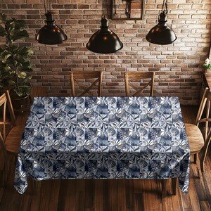 May include: A rectangular tablecloth with a blue and white floral pattern covers a wooden table. The pattern is a repeating design of stylized flowers and geometric shapes. The table is surrounded by wooden chairs, and the background features a brick wall.