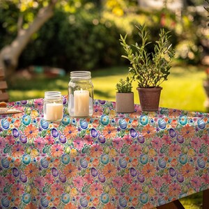 May include: A white tablecloth with a colorful floral pattern featuring pastel-colored eggs and green leaves. The tablecloth is laid out on a wooden table with two glass jars and a plant in a terracotta pot.