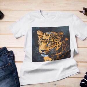 Pode incluir: Camiseta branca com uma estampa gráfica de um rosto de leopardo. O leopardo tem manchas laranja e pretas sobre um fundo escuro.