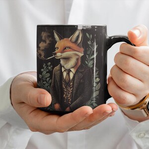 Puede incluir: Una taza de cerámica negra con un gráfico de un zorro con traje y fumando una pipa. El zorro tiene pelaje naranja y un pecho blanco. El fondo es oscuro con follaje verde.