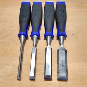 May include: Four chisels with blue and black handles and silver blades are arranged on a wooden surface. The handles have the brand name "Marples" embossed. The blades vary in width and shape, indicating different uses for woodworking.