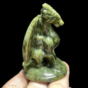 Op de afbeelding: Een gebeeldhouwd groene stenen drakenbeeldje met gedetailleerde vleugels en een zittende pose. De draak is lichtgroen van kleur met donkerdere groene adering. Het beeldje wordt in een hand gehouden tegen een zwarte achtergrond.