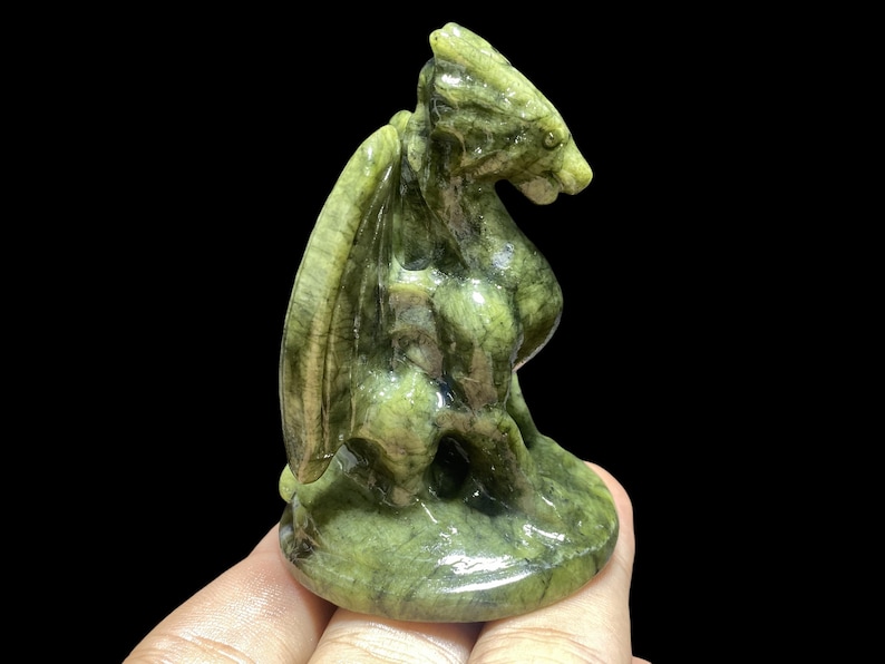 Op de afbeelding: Een gebeeldhouwd groene stenen drakenbeeldje met gedetailleerde vleugels en een zittende pose. De draak is lichtgroen van kleur met donkerdere groene adering. Het beeldje wordt in een hand gehouden tegen een zwarte achtergrond.