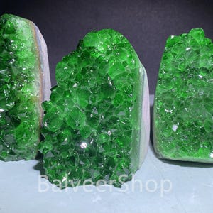 May include: Three vibrant green crystal clusters with a sparkling, faceted surface. The crystals are mounted on a light-colored base, creating a striking contrast. The crystals are a rich, emerald green color.