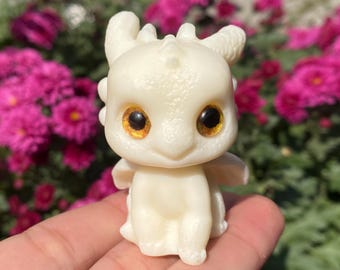 May include: A small, off-white dragon figurine with large, golden-brown eyes. The dragon has small wings and is sitting upright. The background features vibrant pink flowers and green foliage.