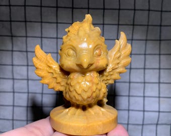 May include: A carved yellow stone figurine of a mythical bird with large eyes, a pointed beak, and outstretched wings. The creature features textured plumage details and stands on a circular base, showcasing a smooth, polished surface finish.