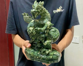 Puede incluir: Una detallada escultura de un dragón verde. El dragón está tallado en piedra con un patrón verde moteado. El dragón está en una pose enrollada, con la cabeza levantada y un pequeño objeto en su garra. La escultura es sostenida por una persona.