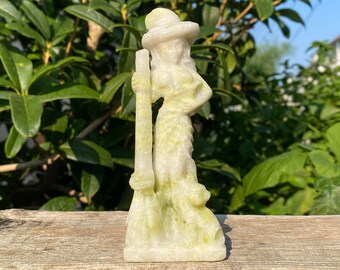 May include: A carved stone figurine of a woman wearing a hat and holding a staff. The sculpture is a pale yellow-green color and stands on a rectangular base. The background is blurred green foliage.