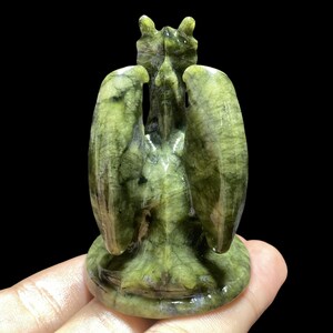 Op de afbeelding: Een groene stenen sculptuur van een gevleugeld wezen met een gedetailleerd ontwerp. De sculptuur heeft een getextureerd oppervlak en staat tegen een zwarte achtergrond. De vleugels zijn gespreid en de figuur heeft een gedetailleerde kop.