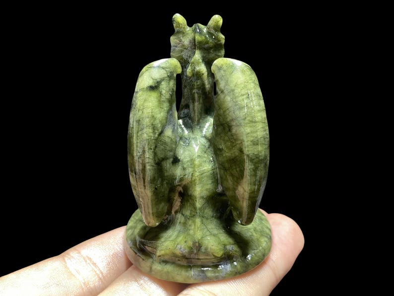 Op de afbeelding: Een groene stenen sculptuur van een gevleugeld wezen met een gedetailleerd ontwerp. De sculptuur heeft een getextureerd oppervlak en staat tegen een zwarte achtergrond. De vleugels zijn gespreid en de figuur heeft een gedetailleerde kop.