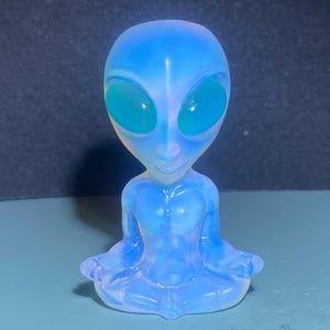 Op de afbeelding: Een lichtblauwe alien figuur met grote, ovale ogen en een serene uitdrukking. De figuur zit in een zittende positie met de handen op de knieën. De figuur heeft een gladde, doorschijnende uitstraling.