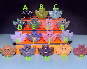 Puede incluir: Una colección de pequeñas figuras de murciélagos talladas en varios colores y materiales. Los murciélagos se exhiben en una superficie escalonada, cada uno etiquetado con una letra. Los colores incluyen negro, verde, rosa y morado.